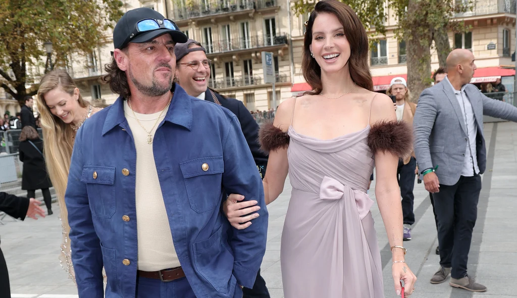 Lana Del Rey z mężem w Paryżu Elegancko ubrana kobieta w długiej, jasnofioletowej sukni z futrzanymi akcentami na ramionach idzie uśmiechnięta, trzymając pod ramię mężczyznę w niebieskiej kurtce i czapce z daszkiem. W tle grupa osób i zabudowa miejska.