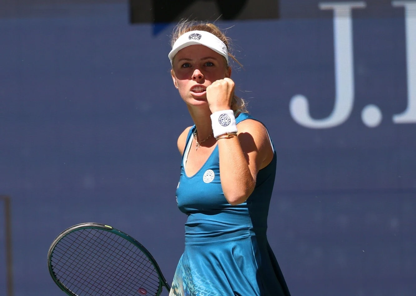 Tenisistka w niebieskiej sukience po udanym zagraniu unosi zaciśniętą pięść w geście triumfu, trzymając rakietę w drugiej ręce i nosząc białą opaskę oraz daszek sportowy.