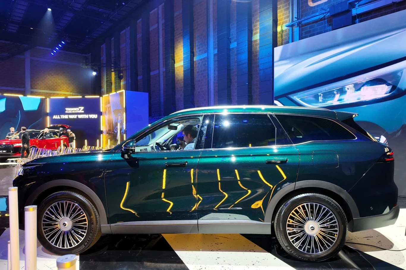 Luksusowy zielony SUV na wystawie motoryzacyjnej, oświetlony kolorowymi światłami i otoczony nowoczesną scenografią z neonowymi refleksami na karoserii.