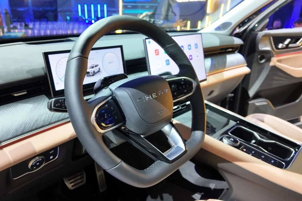 Wewnątrz nie znajdziemy zbyt wiele fizycznych przełączników. Kilka zostało na konsoli środkowej. Obok znajduje się indukcyjna ładowarka. Nowoczesna deska rozdzielcza samochodu osobowego z cyfrowymi wyświetlaczami i kierownicą marki Chery, dotykowy ekran centralny i eleganckie wykończenie wnętrza.