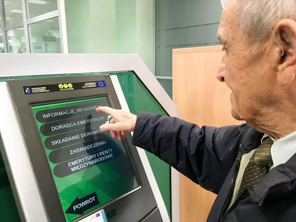 Nie korzystasz z aplikacji na smartfonie? Nadal możesz załatwić sprawy w oddziale ZUS Starszy mężczyzna korzysta z dotykowego terminala informacyjnego w urzędzie, wybierając jedną z dostępnych opcji na ekranie dotyczącą emerytur lub świadczeń społecznych.