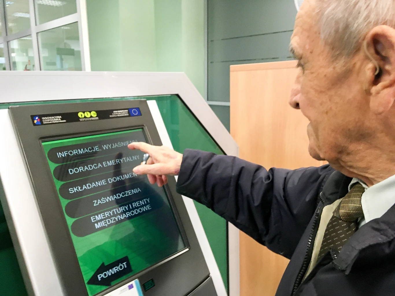 Nie korzystasz z aplikacji na smartfonie? Nadal możesz załatwić sprawy w oddziale ZUS Starszy mężczyzna korzysta z dotykowego terminala informacyjnego w urzędzie, wybierając jedną z dostępnych opcji na ekranie dotyczącą emerytur lub świadczeń społecznych.