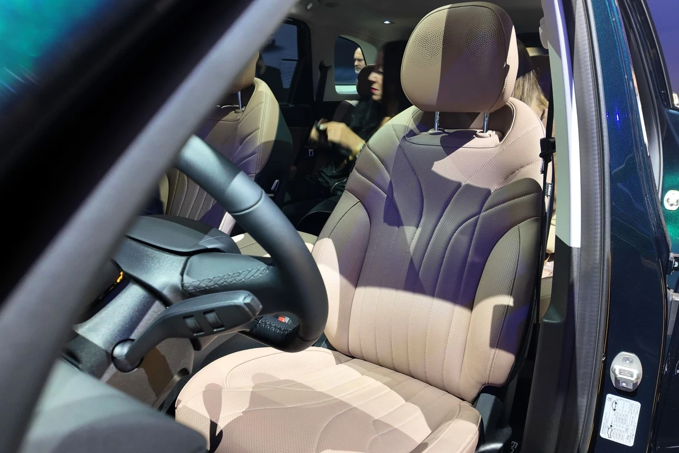 Przedni fotel samochodu z jasną tapicerką, kierownica oraz fragment deski rozdzielczej, w tle widoczne dwie kobiety siedzące na tylnej kanapie.