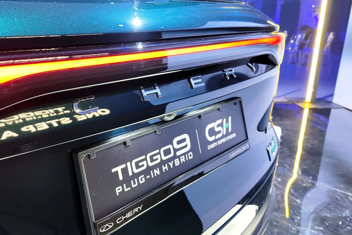 Tylna część samochodu osobowego marki Chery z widocznym oznaczeniem modelu Tiggo 9 Plug-in Hybrid, nowoczesny design świateł LED, czarna tablica rejestracyjna ekspozycyjna, otoczenie oświetlone delikatnie niebieskim światłem.