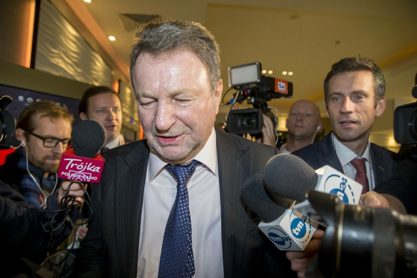 Mężczyzna w garniturze otoczony przez dziennikarzy i kamery, na pierwszym planie mikrofony z logotypami różnych mediów, scena oświetlona światłem sztucznym, tło rozmyte, z obecnością kilku osób w garniturach