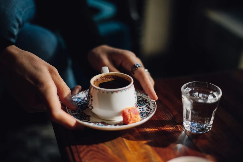 Kawa "po turecku" ma wielu zwolenników, ale niesie za sobą pewne zagrożenia Dłoń sięga po filiżankę kawy ustawioną na ozdobnym spodku z kawałkiem lokum oraz szklanką wody na drewnianym stole.
