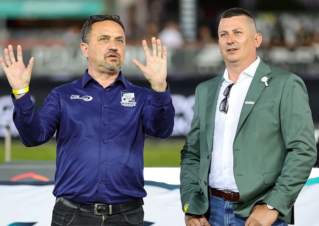 Były już prezes Włókniarza Michał Świącik i Bartłomiej Januszko, właściciel Krono-Plastu. Dwóch mężczyzn w średnim wieku stoi obok siebie, jeden gestykuluje uniesionymi dłońmi, obaj są elegancko ubrani w koszule i marynarki, w tle rozmyty tłum oraz elementy wydarzenia sportowego.