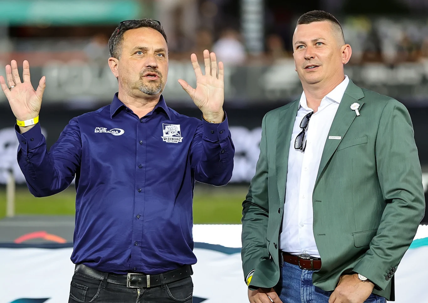 Dwóch mężczyzn w średnim wieku stoi obok siebie, jeden gestykuluje uniesionymi dłońmi, obaj są elegancko ubrani w koszule i marynarki, w tle rozmyty tłum oraz elementy wydarzenia sportowego.