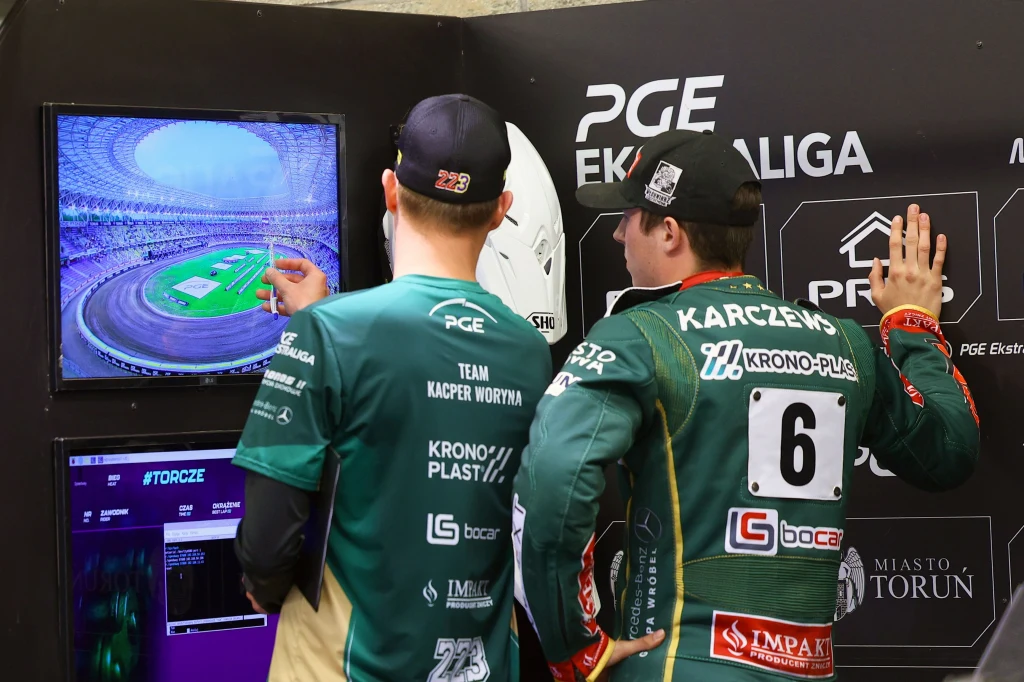 Franciszek Karczewski analizuje geometrię toru. Od jego postawy zależy los Włókniarza. Dwóch żużlowców w zielonych kombinezonach analizuje na monitorze obraz stadionu żużlowego, zwracając uwagę na szczegóły toru.