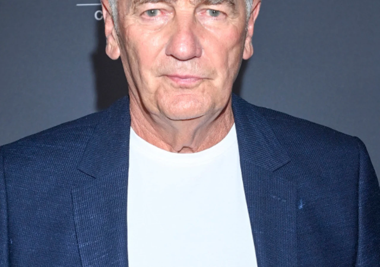 Karol Strasburger Starszy mężczyzna o krótkich, siwych włosach, ubrany w granatową marynarkę i białą koszulkę, patrzy prosto w obiektyw na tle ciemnej, jednolitej ściany.