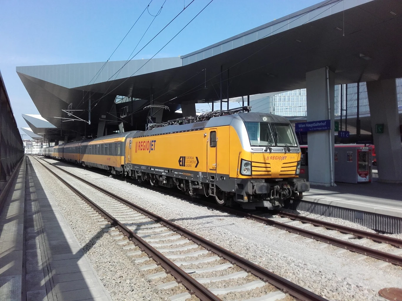 RegioJet pojedzie do kolejnych miast w Polsce. W planach Wrocław. Nowoczesny żółty pociąg pasażerski Leo Express stojący przy peronie dużego, zadaszonego dworca kolejowego, po lewej stronie widać przeszkloną barierę a w tle budynki miejskie.