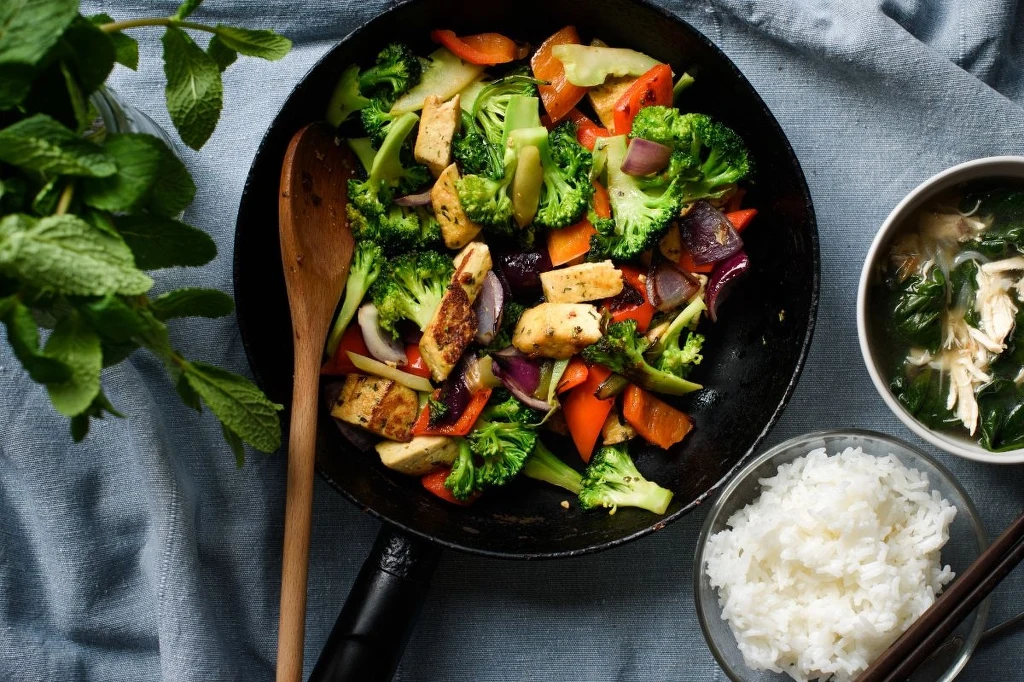 Tę sałatkę możesz jeść z komosą i kaszą z boku lub wymieszane z warzywami. W obu wersjach smakuje świetnie Kolorowa mieszanka warzyw smażonych na patelni z tofu, po lewej świeże zioła, po prawej miseczka z białym ryżem oraz zupa z kurczakiem i warzywami, całość na niebieskiej tkaninie.