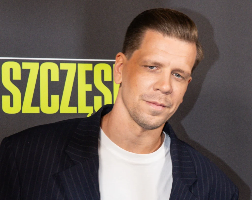 Wojciech Szczęsny na premierze filmu dokumentalnego "Szczęsny" Mężczyzna w jasnej koszulce i granatowej marynarce pozuje na tle czarnej ścianki z żółtym napisem.