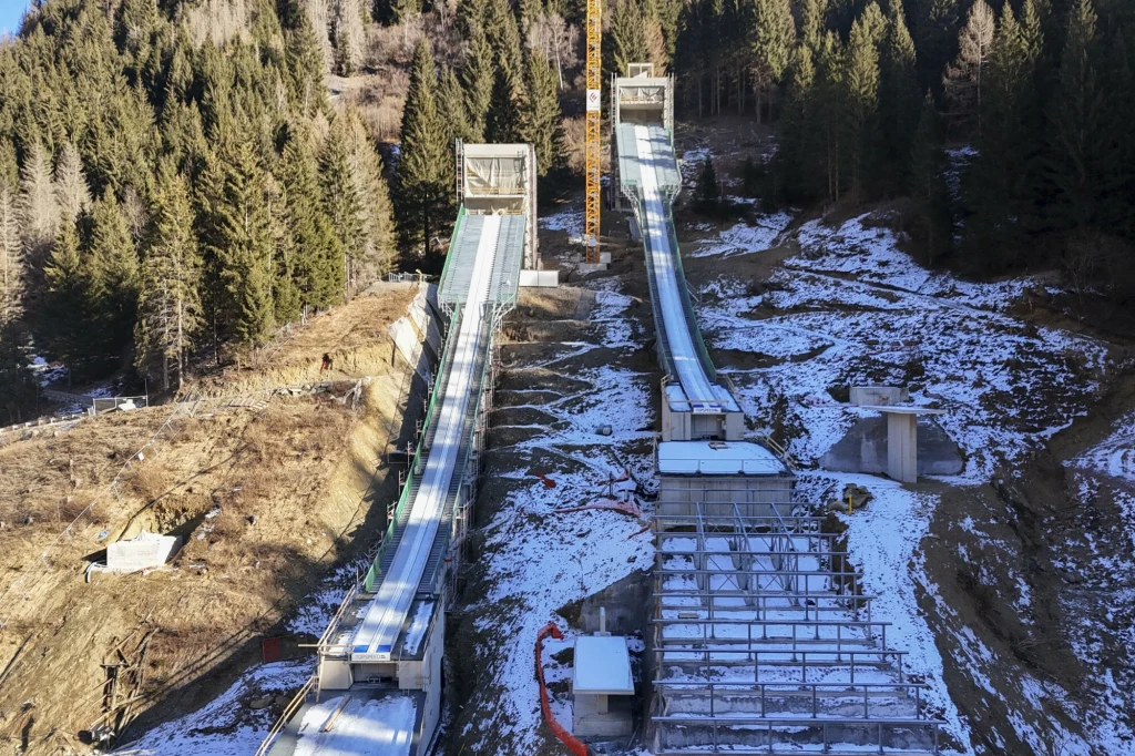 Skocznie narciarskie w Predazzo zostały przebudowane specjalnie na ZIO 2026 Kompleks skoczni narciarskich położony na tle wzgórza z częściowo ośnieżonym stokiem, otoczony gęstym lasem świerkowym, widoczne konstrukcje techniczne oraz przygotowane lądowisko skoczni.