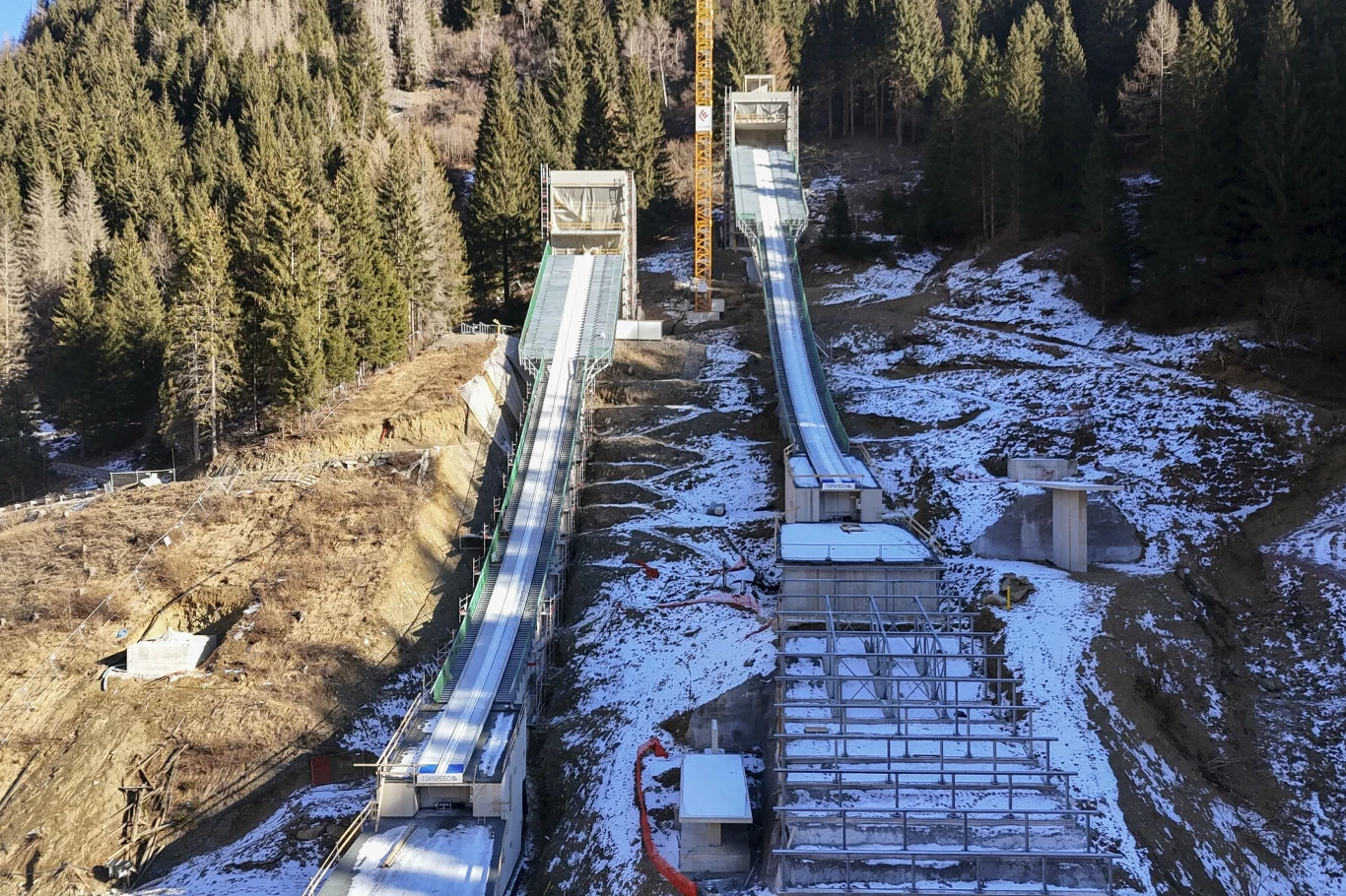 Kompleks skoczni narciarskich położony na tle wzgórza z częściowo ośnieżonym stokiem, otoczony gęstym lasem świerkowym, widoczne konstrukcje techniczne oraz przygotowane lądowisko skoczni.