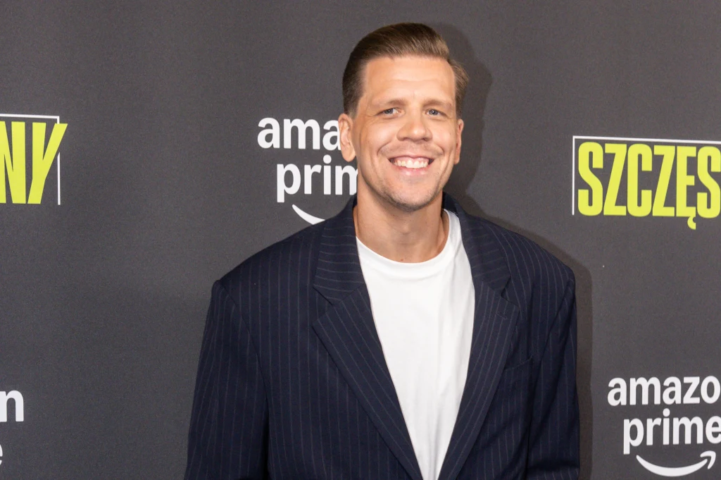 Wojciech Szczęsny Uśmiechnięty mężczyzna w garniturze w pionowe paski oraz białym T-shircie, stojący na tle czarnej ścianki z logotypami amazon prime i napisem SZCZĘŚ.