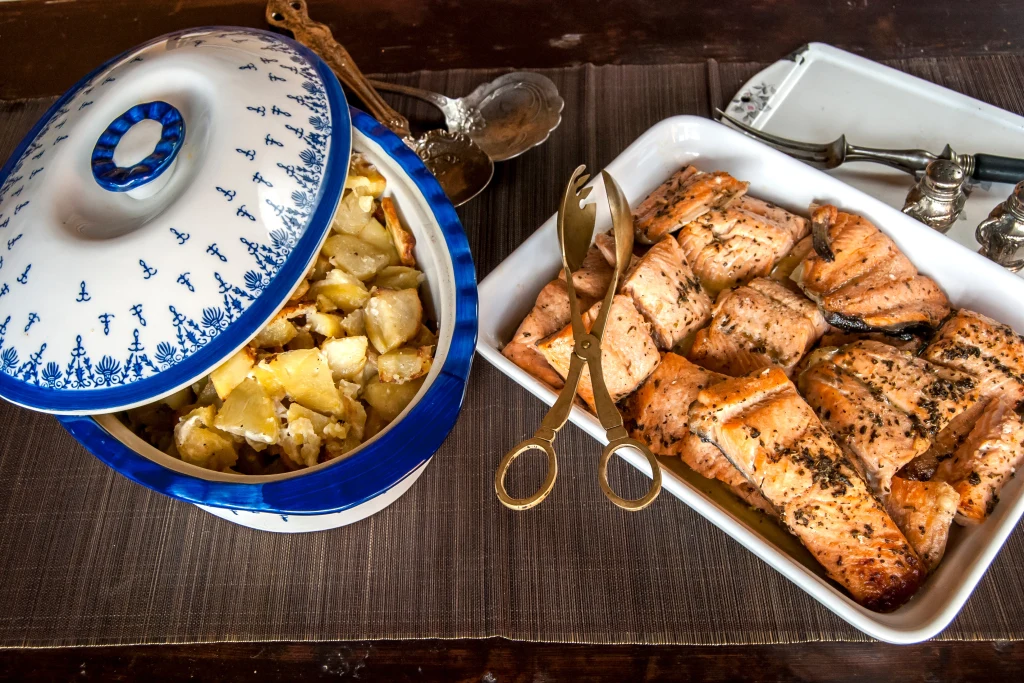W diecie kobiet w wieku okołomenopauzalnym, istotną rolę ogrywają kwasy omega-3. Ich źródłem są m.in. tłuste ryby morskie Rumiane, pieczone filety z łososia przyprawione ziołami w białym naczyniu oraz ziemniaki w porcelanowej misie z niebieskim wzorem, obok sztućce do podawania potraw.