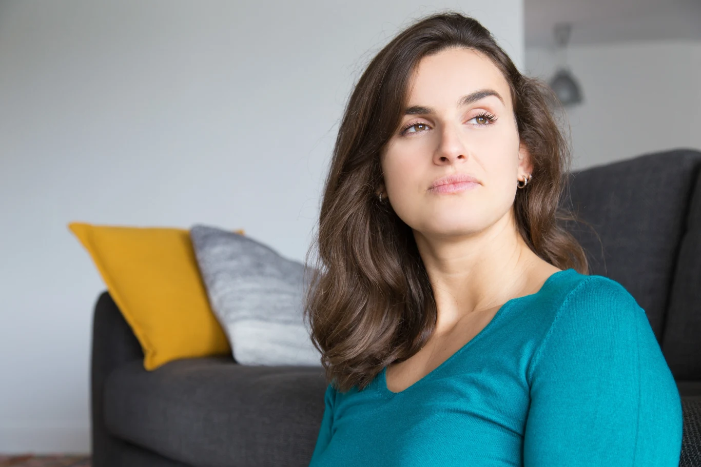 Młoda kobieta z długimi, brązowymi włosami siedzi w salonie i patrzy w bok, uśmiechając się lekko, w tle szara sofa z żółtą i szarą poduszką.