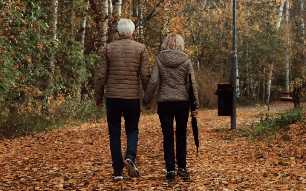 Regularna aktywność fizyczna i redukowanie stresu pomogą obniżyć wiek metaboliczny Para starszych osób spaceruje alejką w parku otoczoną jesiennymi liśćmi, trzymając się za ręce i idąc w stronę gęstego lasu.
