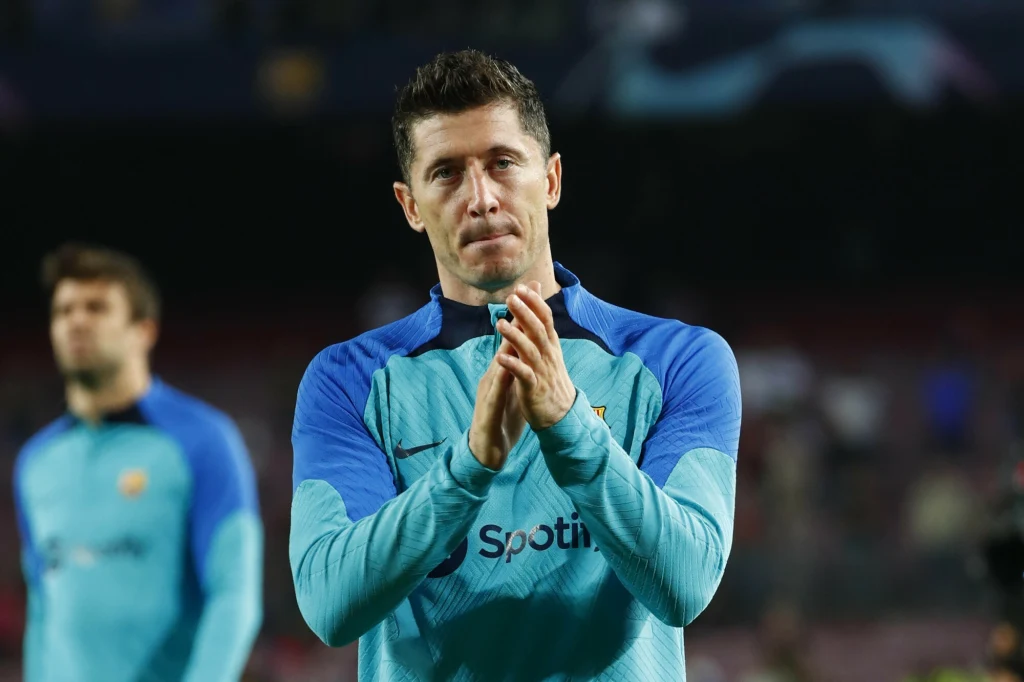 Robert Lewandowski Piłkarz w sportowej koszulce z napisem 'Spotify', składa dłonie jak do oklasków, patrząc w stronę obiektywu, w tle rozmyta postać na stadionie.