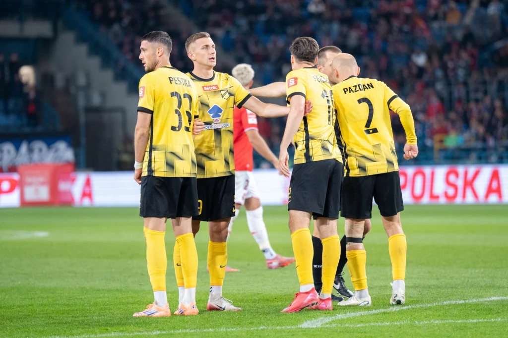 Grupa piłkarzy w żółto-czarnych strojach stoi razem na murawie stadionu, wyrażając emocje podczas meczu, w tle widownia i fragment reklamy.