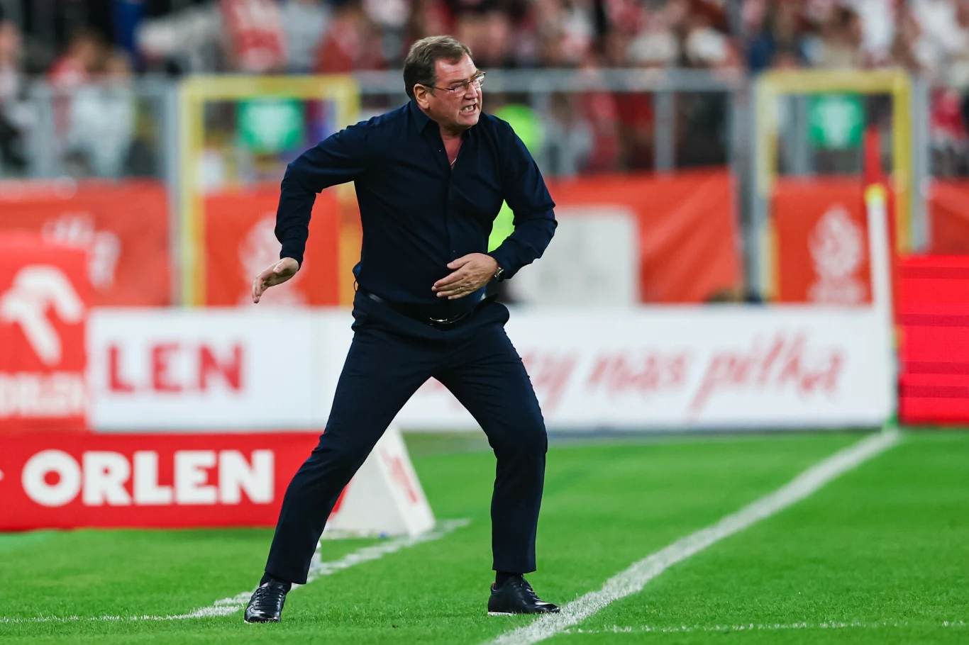 Jn Urban podczas meczu z Finlandią Mężczyzna w średnim wieku w ciemnym garniturze gestykuluje energicznie na zielonym boisku piłkarskim, w tle rozmazana widownia oraz reklamy przy linii bocznej.