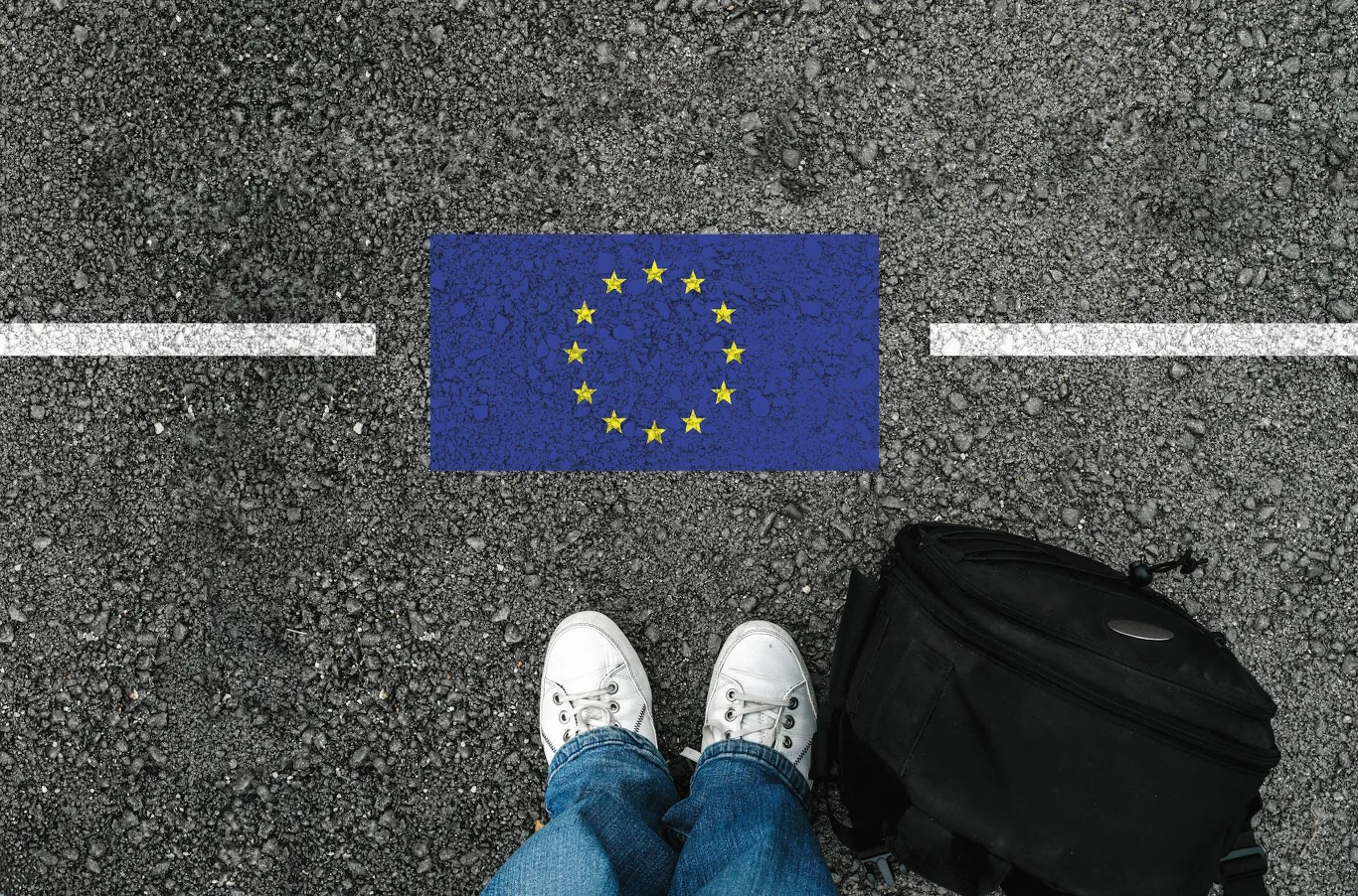 Para białych butów i plecak na asfalcie przy namalowanej fladze Unii Europejskiej oddzielonej białą linią.