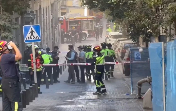 W Madrycie zwalił się budynek. Na miejscu trwa akcja służb Służby ratunkowe oraz policjanci zabezpieczają teren na ulicy, otoczony taśmą, za którą znajduje się lekko zadymiona strefa. W tle widoczny wóz strażacki oraz samochody. Pracownicy są wyposażeni w odzież ochronną i kaski.