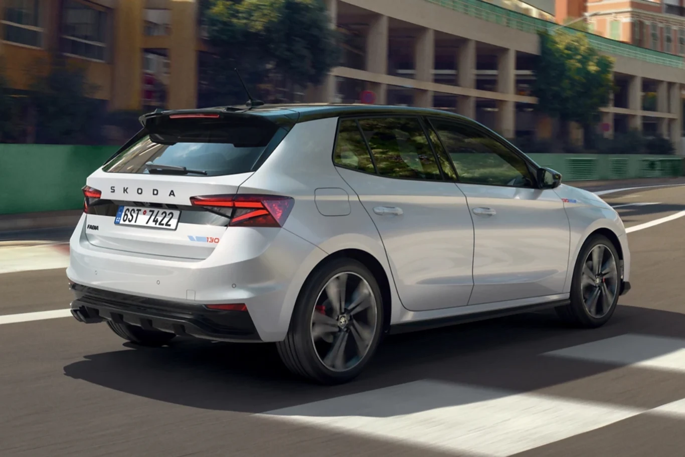 Srebrny samochód marki Skoda porusza się po miejskiej drodze, w tle widoczne są zabudowania oraz zieleń miejska.