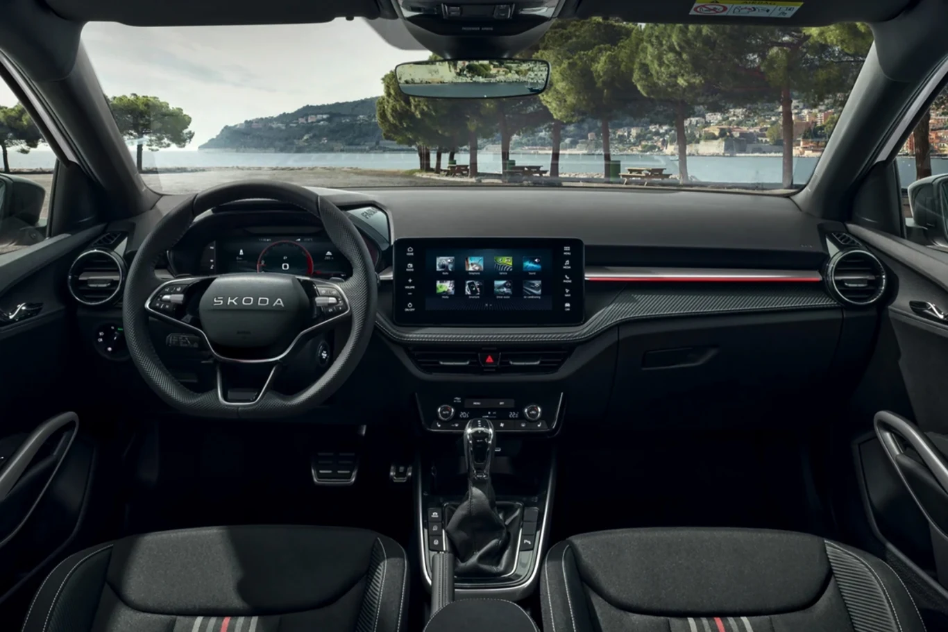 Nowoczesna deska rozdzielcza samochodu marki Skoda z cyfrowym wyświetlaczem, dużym ekranem dotykowym pośrodku, sportową kierownicą, konsolą środkową oraz widokiem na część krajobrazu przez przednią szybę.