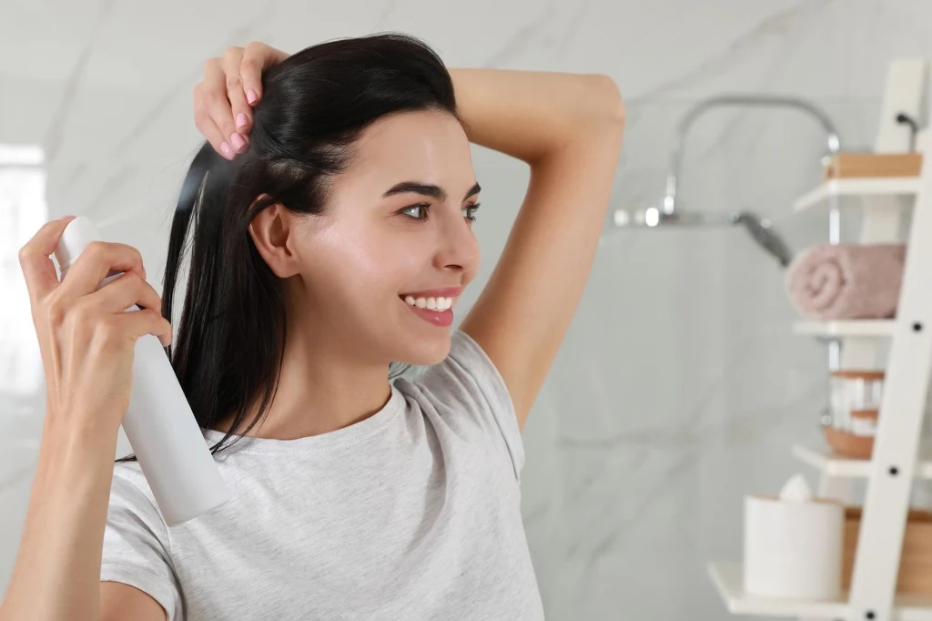 Aerozole takie jak lakier do włosów powinny być przechowywane poza łazienką Uśmiechnięta kobieta z ciemnymi włosami trzymająca w jednej ręce butelkę ze sprayem i rozprowadzająca produkt na włosach, w tle jasna łazienka z regałem i ręcznikami.