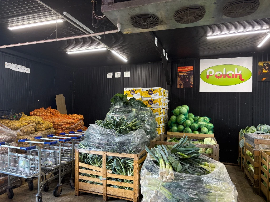 Sprzedawca: Kontrahenci skarżą się na nadprodukcję ziemniaków Wnętrze warzywniaka z regałami wypełnionymi świeżymi warzywami, takimi jak ziemniaki, cebula, arbuzy, pory i zielone warzywa liściaste; w tle widoczna reklama ze słowem 'Polak'.