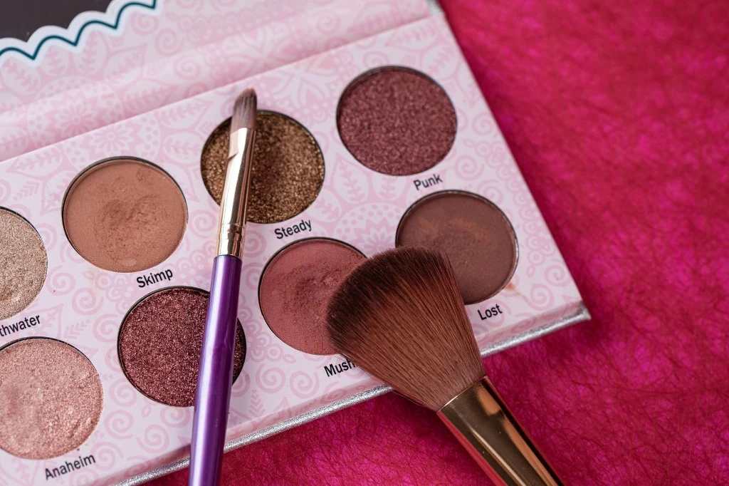 Cienie do powiek, pudry i bronzery źle zniosą wilgoć w łazience Paleta cieni do powiek w odcieniach brązu i różu z dwoma pędzlami do makijażu na tle różowego materiału.