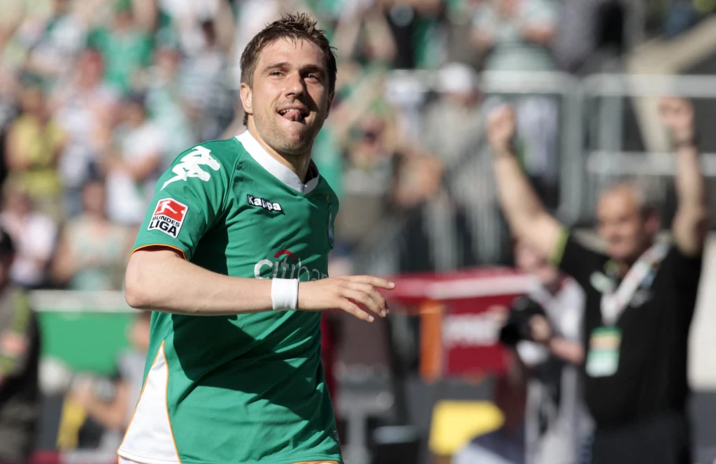 Ivan Klasnić Piłkarz w zielonym stroju podczas meczu, na tle kibiców na trybunach, z radosnym wyrazem twarzy po udanej akcji sportowej.