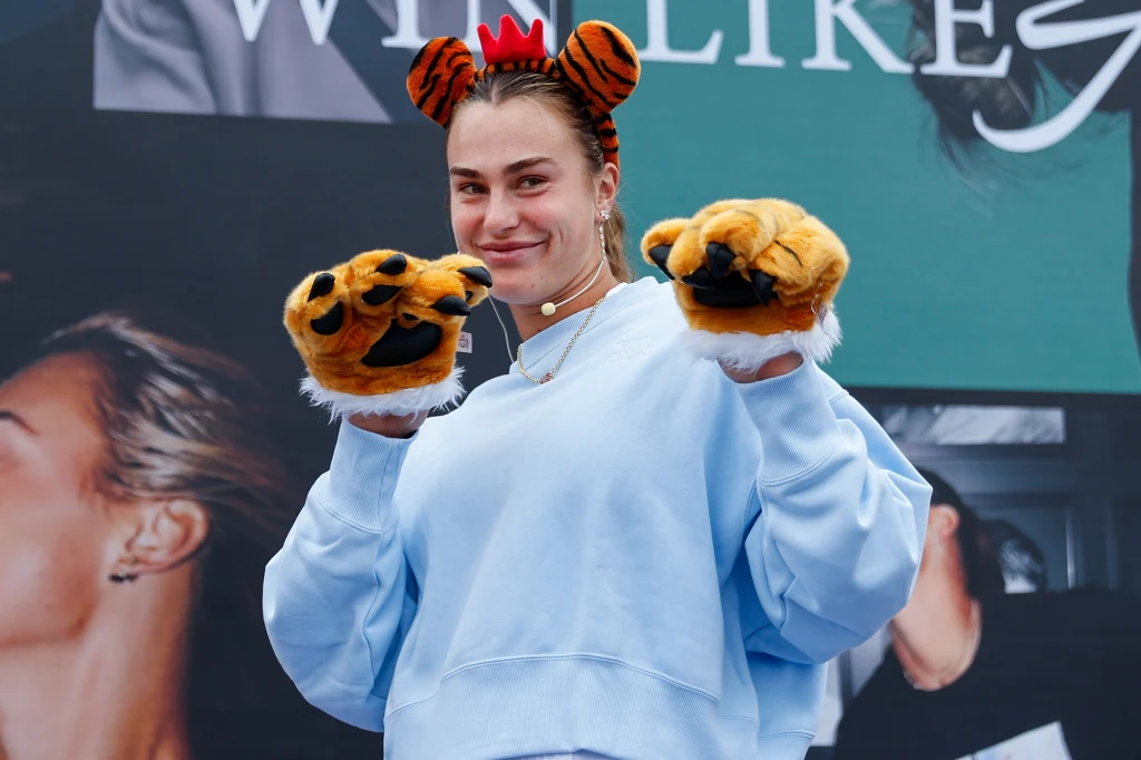 Aryna Sabalenka w Wuhan Kobieta w niebieskiej bluzie pozuje z uśmiechem, mając na głowie opaskę z uszami tygrysa i zakładając na ręce pluszowe łapy przypominające łapy tygrysa. W tle widoczny duży plakat ze zdjęciami.