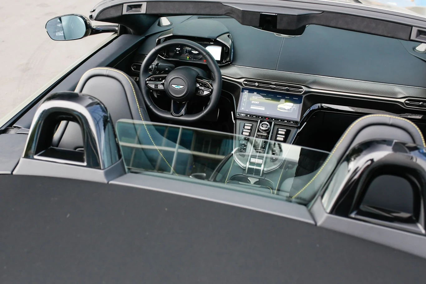 Automatyczny dach Astona Martina Vantage'a Roadstera można złożyć i rozłożyć w 6,8 sekundy. Możemy to zrobić w czasie jazdy z prędkością do 50 km/h. Wnętrze luksusowego samochodu kabrioletu z widocznym nowoczesnym panelem sterowania, kierownicą oraz wykończeniem ze skóry i metalu, ujęcie wykonane zza foteli.