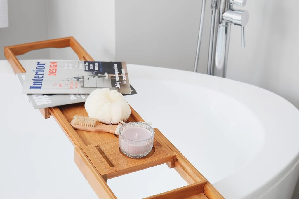 Czasopisma i książki w łazience? To kiepski pomysł Bambusowa półka na wannę z rozłożonymi na niej magazynami wnętrzarskimi, gąbką, szczotką do masażu oraz zapaloną świecą, w tle biała, nowoczesna wanna i bateria łazienkowa.