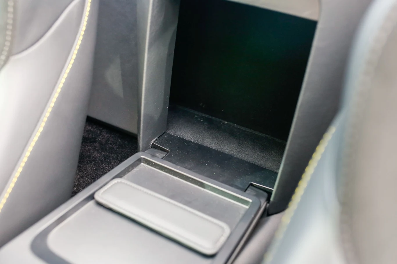 Trudno powiedzieć, co pomieści ten płytki schowek. Nie ma tutaj przejścia do bagażnika. otwarty schowek znajdujący się między siedzeniami samochodu, wykonany z czarnego tworzywa, z widocznymi przeszyciami na bocznych krawędziach siedzeń