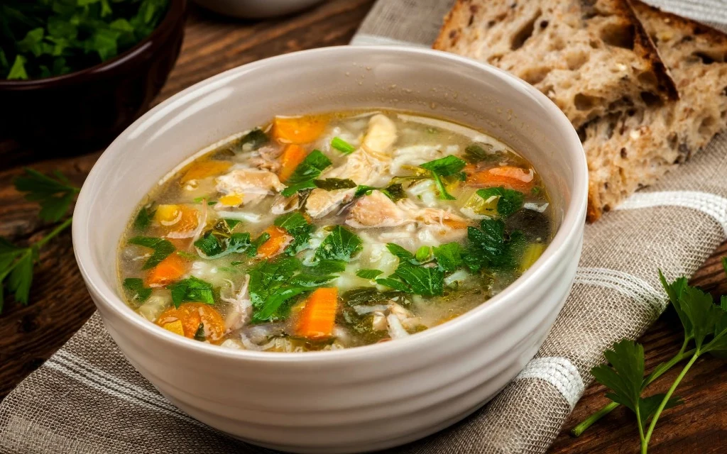 Zjedz zupę warzywną na kolację, a sen będzie nieprzerwany Miseczka gorącej zupy z kawałkami marchewki, mięsa z kurczaka, świeżymi ziołami oraz dodatkiem pokrojonego chleba na lnianej serwecie, całość utrzymana w domowej atmosferze.