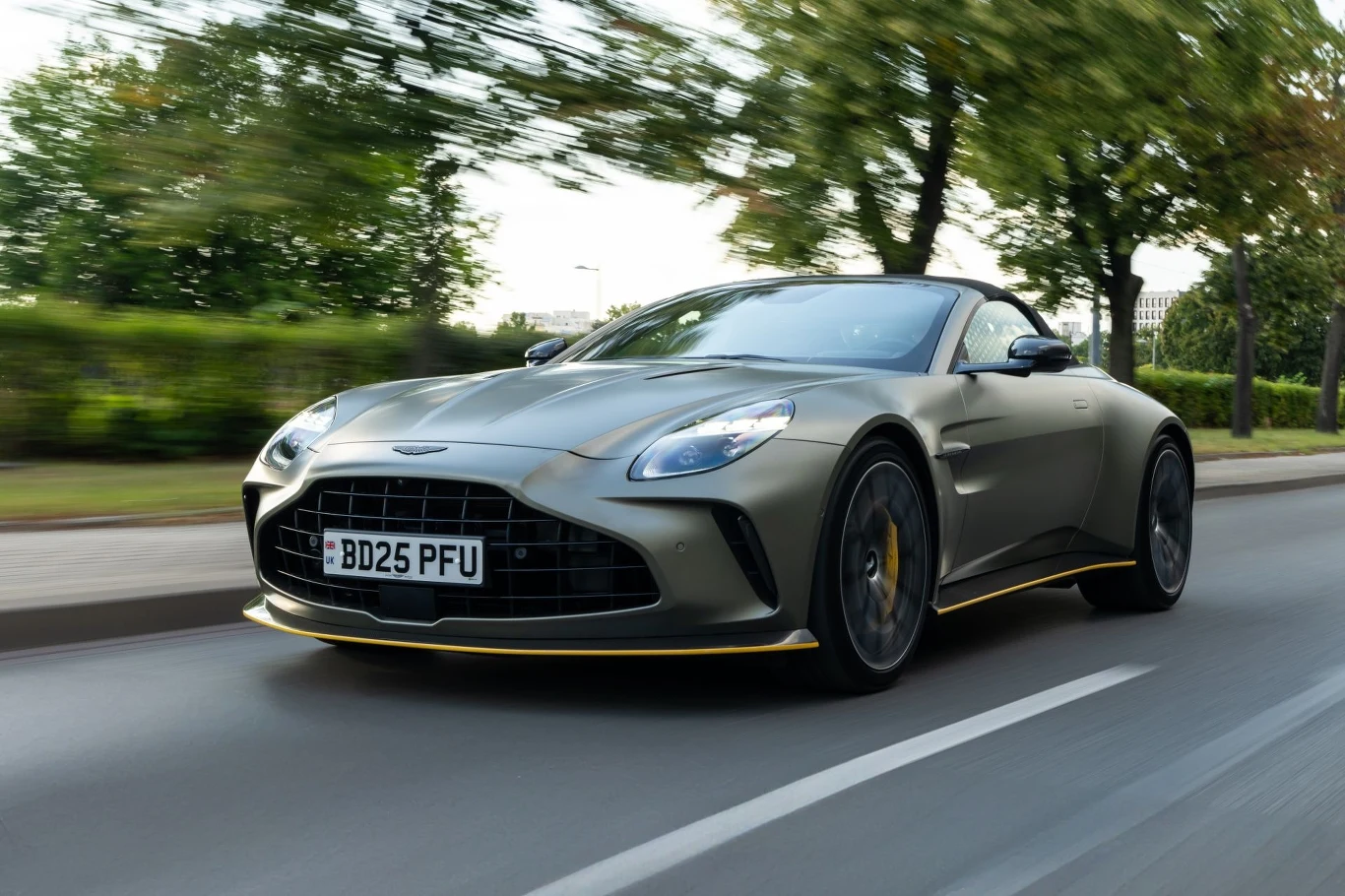 Aston Martin Vantage Roadster to nie jest tylko wersja Coupe z rozkładanym dachem. Oba warianty nadwozia opracowano równolegle. Luksusowy sportowy samochód marki Aston Martin porusza się po miejskiej ulicy, otoczony zielenią i lekko rozmazanym tłem, co sugeruje dynamiczną jazdę.