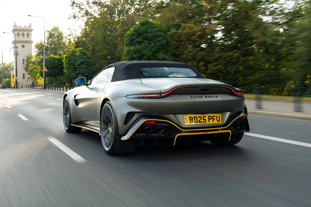 Srebrny samochód sportowy Aston Martin z otwartym dachem jedzie pustą ulicą miejską, otoczony zielenią i niskimi barierkami metalowymi, w tle wieża zegarowa i poranne światło