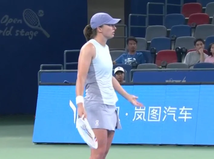 Iga Świątek Zawodniczka tenisowa w sportowym stroju stoi na korcie twardym, trzymając rakietę w dłoni, z wyrazem zdziwienia lub niezadowolenia na twarzy, w tle trybuny z kibicami oraz niebieskie banery reklamowe.