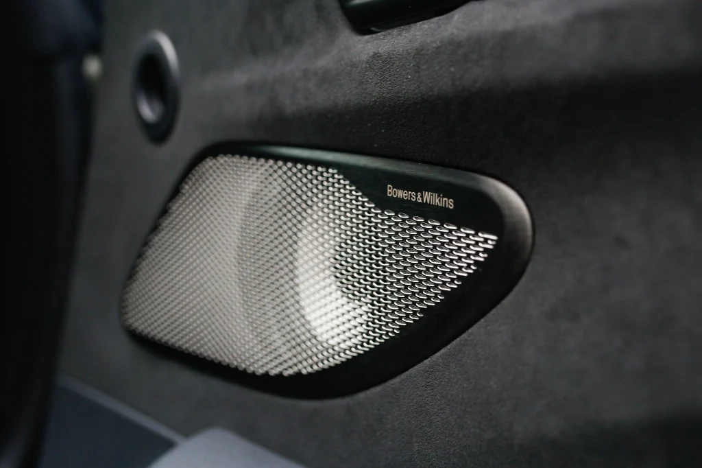Srebrny głośnik Bowers & Wilkins zamontowany w tapicerce drzwi samochodu, z metalową siatką ochronną i logo producenta widocznym po prawej stronie.