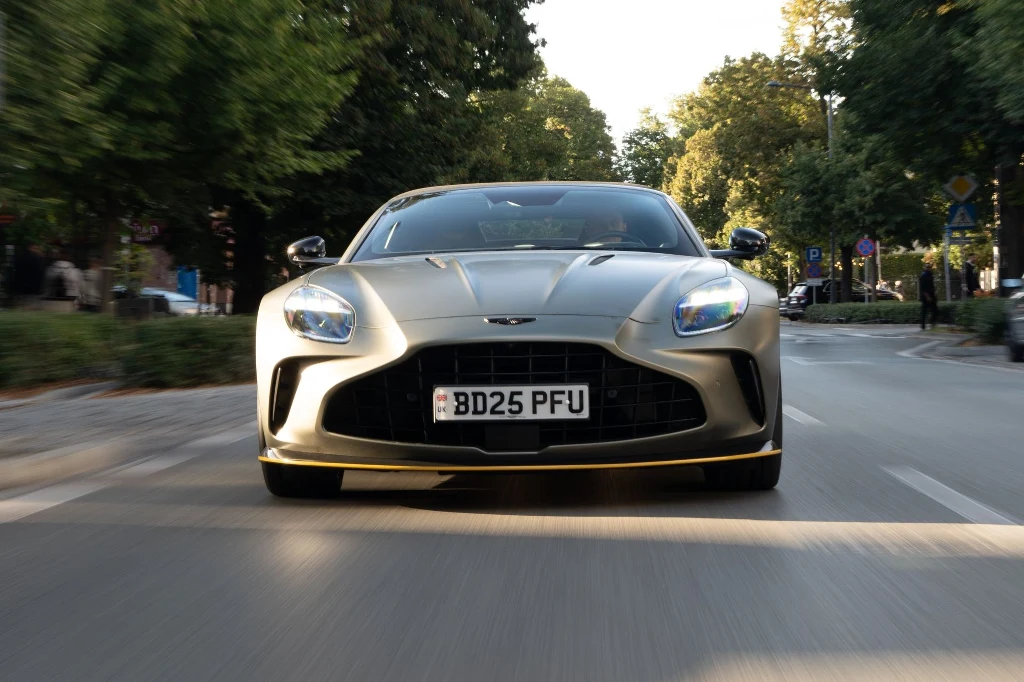 Sportowy samochód marki Aston Martin jedzie ulicą miejską w otoczeniu zielonych drzew i znaków drogowych, widoczny jest z przodu z nisko ustawioną kamerą akcentującą jego dynamiczny charakter.