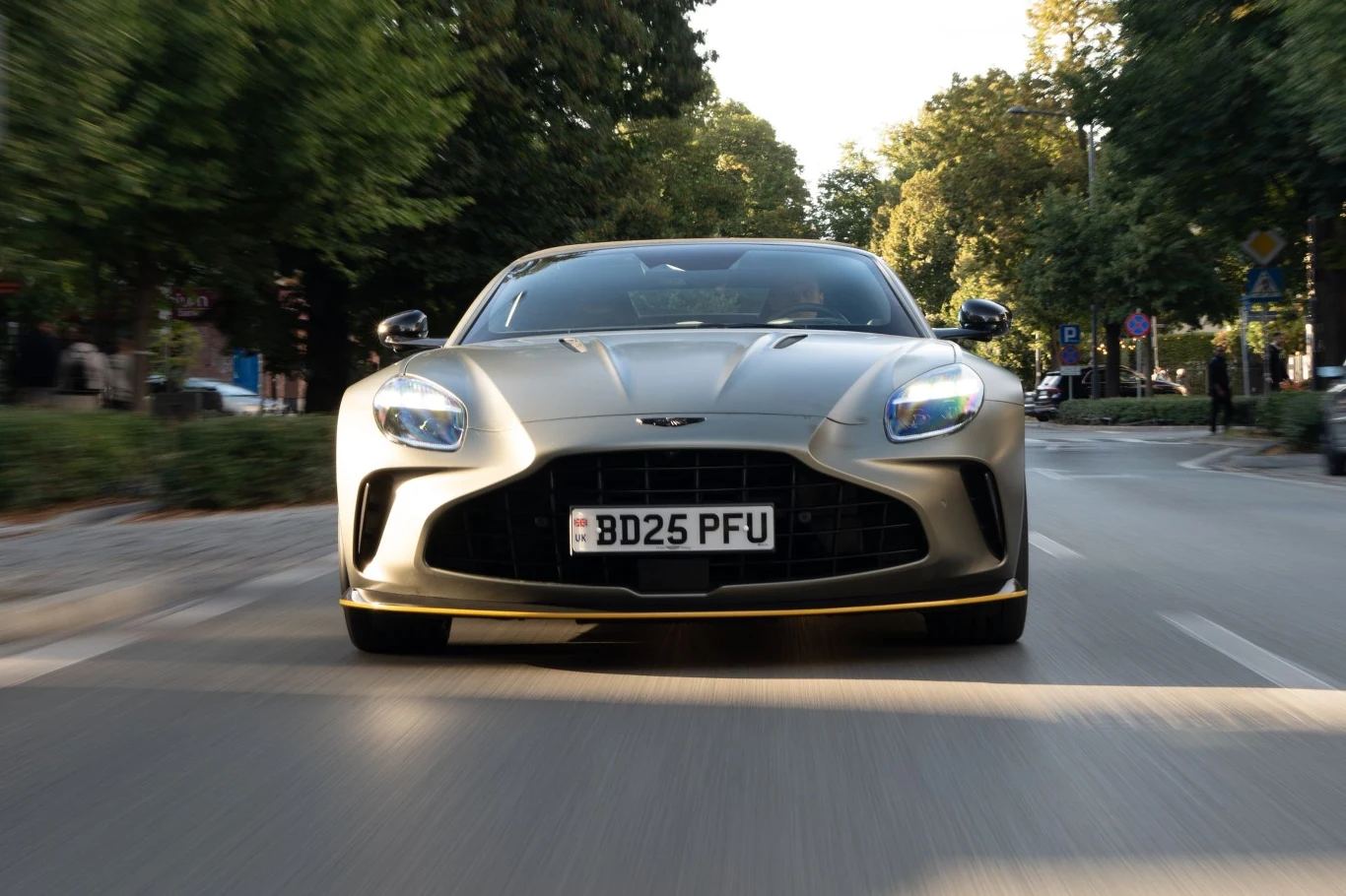 Aston Martin Vantage Roadster jest tylko o 60 kg cięższy od wariantu Coupe. Sportowy samochód marki Aston Martin jedzie ulicą miejską w otoczeniu zielonych drzew i znaków drogowych, widoczny jest z przodu z nisko ustawioną kamerą akcentującą jego dynamiczny charakter.