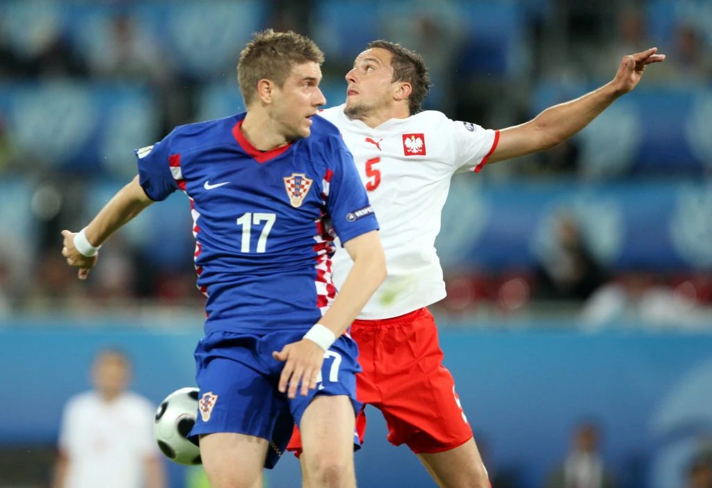 Ivan Klasnić w meczu z Polską na Euro 2008 Dwóch piłkarzy w trakcie walki o piłkę podczas meczu piłkarskiego, jeden ubrany w niebieski strój z numerem 17, drugi w biało-czerwony strój z numerem 5, dynamiczny moment starcia na tle stadionu.