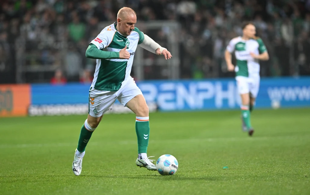 Ivan Klasnić w marcu 2025 roku Piłkarz w zielono-białym stroju biegnie z piłką po murawie podczas meczu piłkarskiego, w tle widoczni są inni zawodnicy oraz kibice na trybunach.