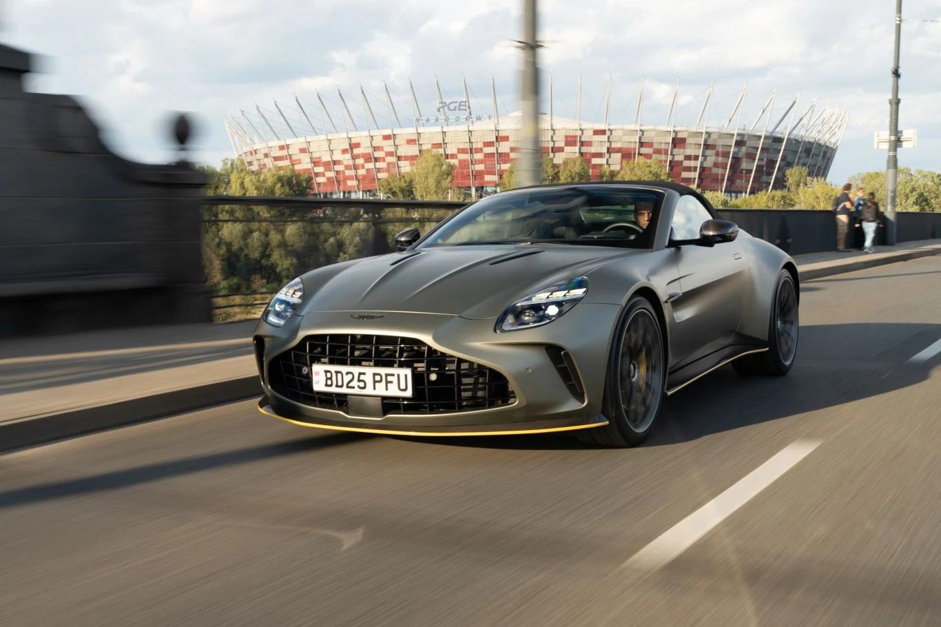 Rozkład masy Astona Martina Vantage'a Roadstera wynosi 49:51. Sportowy samochód w kolorze szarym z widocznymi reflektorami LED jedzie po mieście na tle nowoczesnego stadionu narodowego.