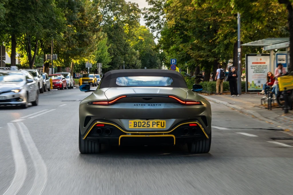 Luksusowy samochód sportowy Aston Martin w kolorze zielonym z widoczną tablicą rejestracyjną jedzie miejską ulicą otoczoną zielonymi drzewami, w tle inne pojazdy oraz przystanek z oczekującymi osobami.