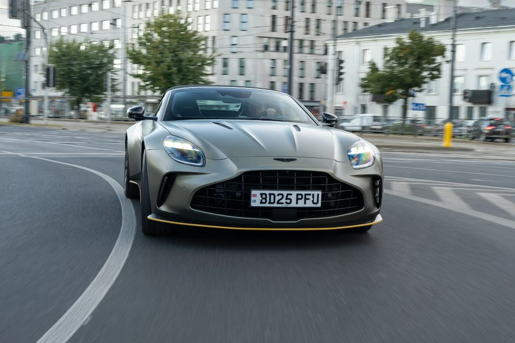 Luksusowy samochód sportowy Aston Martin DB12 w szarym kolorze jedzie po asfaltowej drodze miejskiej, w tle nowoczesne budynki i zielone drzewa.
